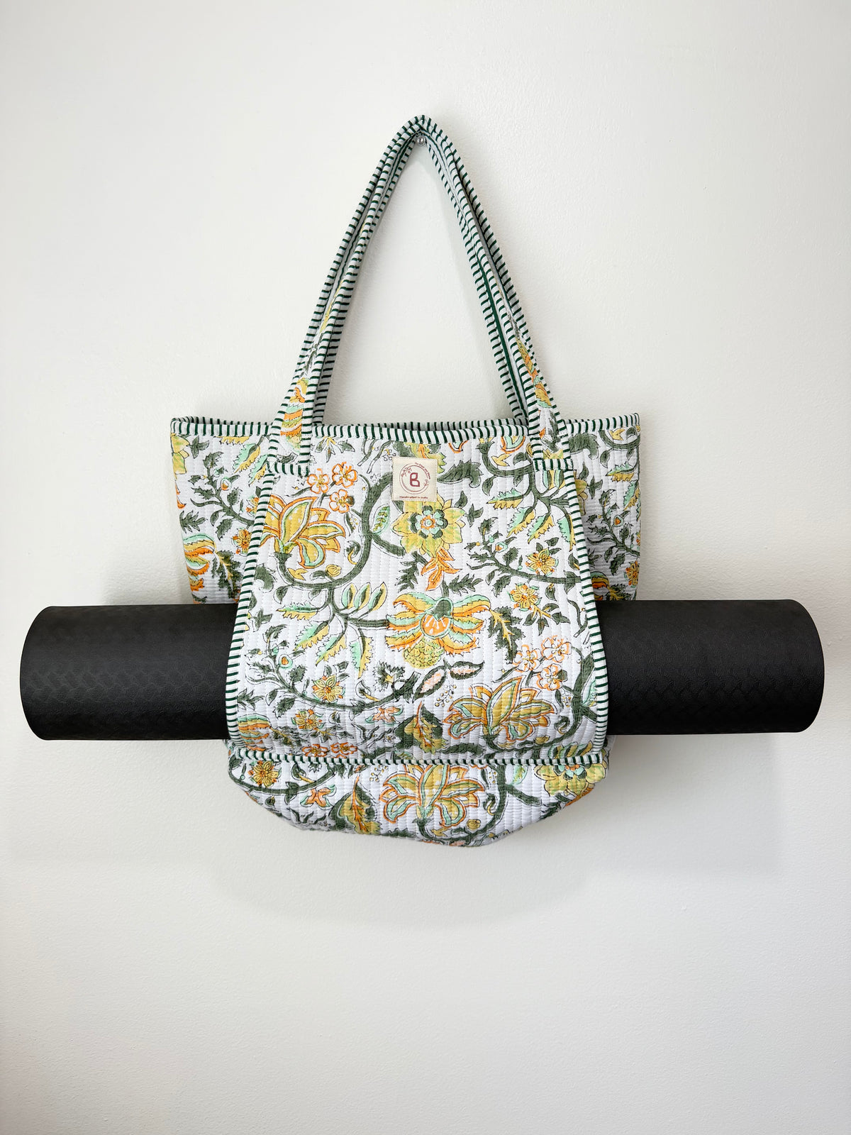 A fabric tote bag with a green, yellow, and white floral pattern, hanging from a black rolled-up yoga mat against a plain white wall.
