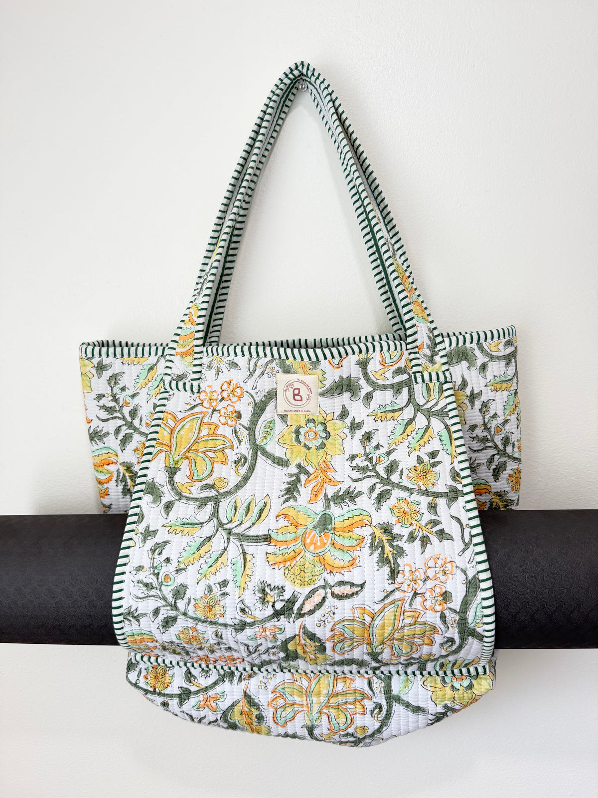 A fabric tote bag with a white background and a colorful floral pattern in yellow, orange, and green, featuring striped green and white shoulder straps and a small circular 'B' logo patch on the front.