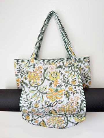 A fabric tote bag with a white background and a colorful floral pattern in yellow, orange, and green, featuring striped green and white shoulder straps and a small circular 'B' logo patch on the front.