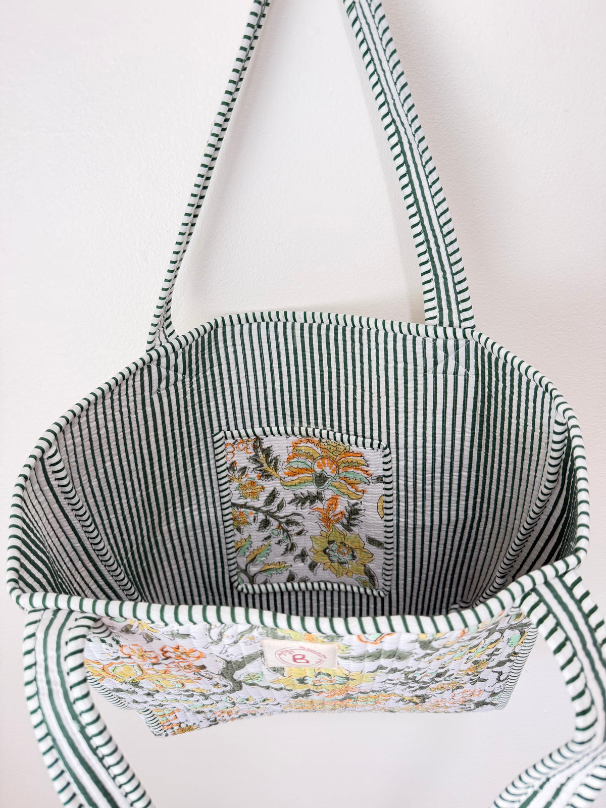 A fabric tote bag with dark green and white vertical stripes on the main body and handles, featuring a front pocket with a white background and a colorful floral print.