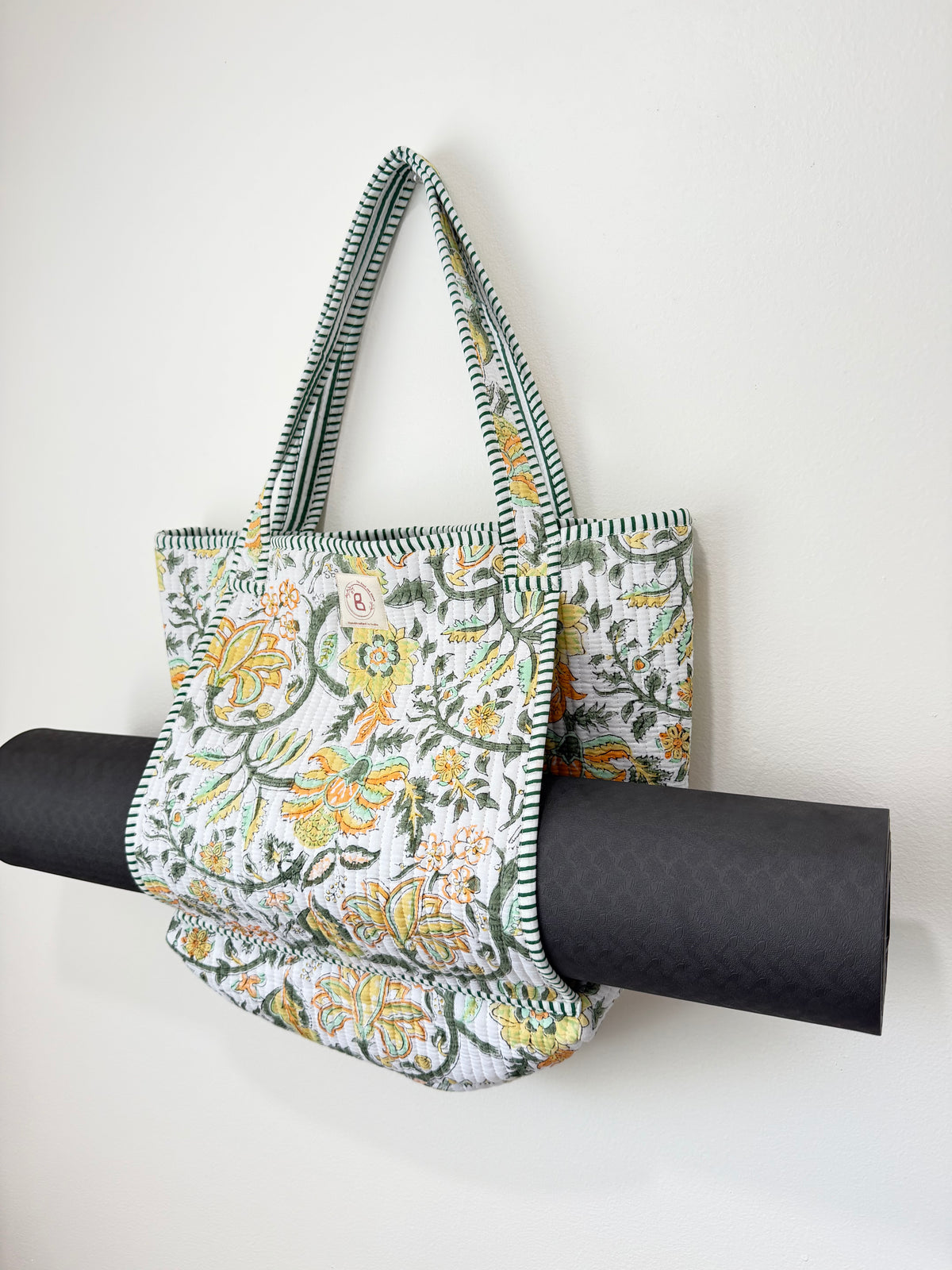 A fabric tote bag with a green, yellow, and white floral pattern hangs against a plain white wall, draped over a rolled black exercise or yoga mat.