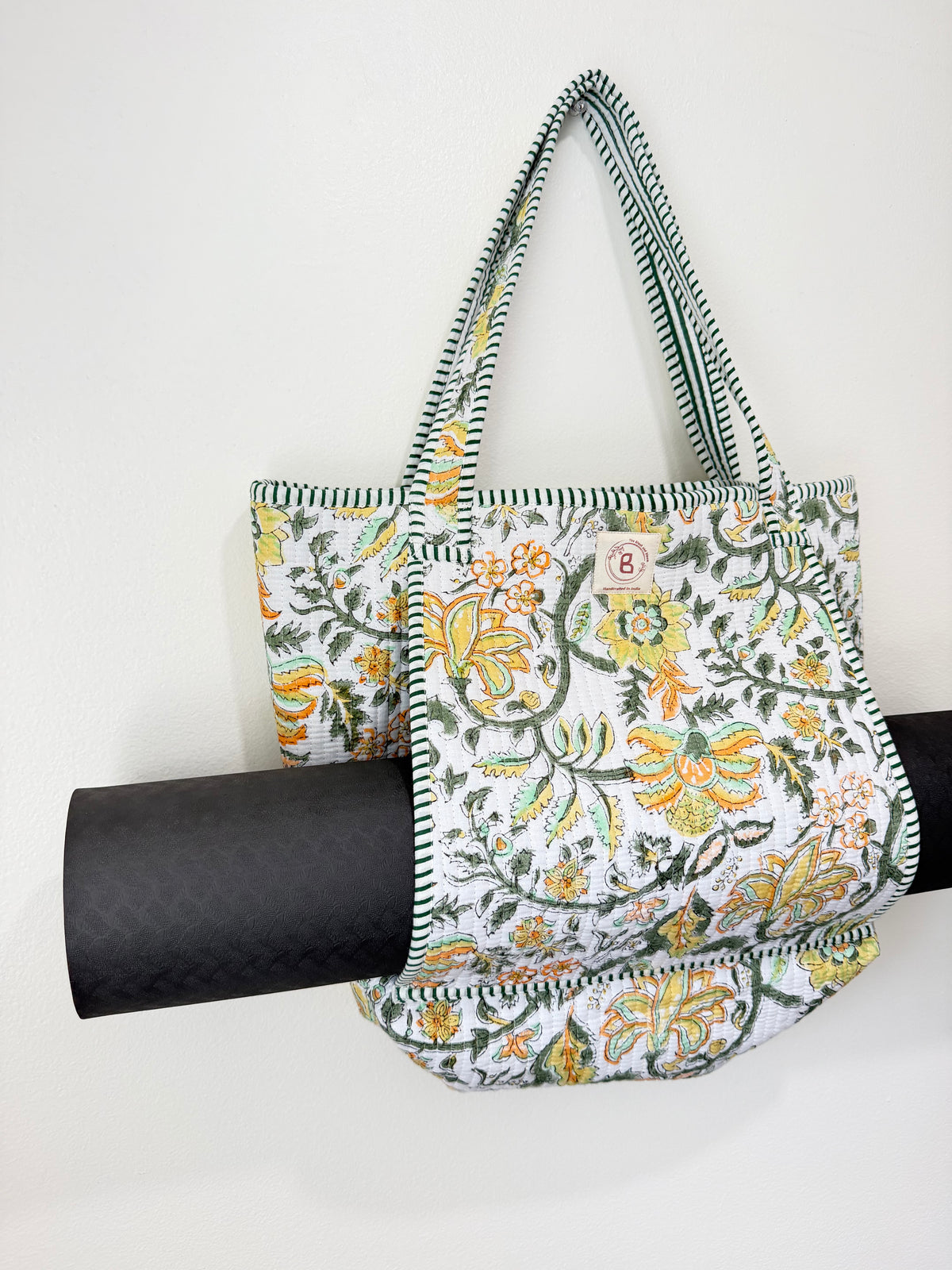A floral tote bag with green and white striped handles, featuring a white background with yellow and green botanical patterns. A black rolled-up yoga mat is draped behind the bag as a prop, set against a plain white wall.