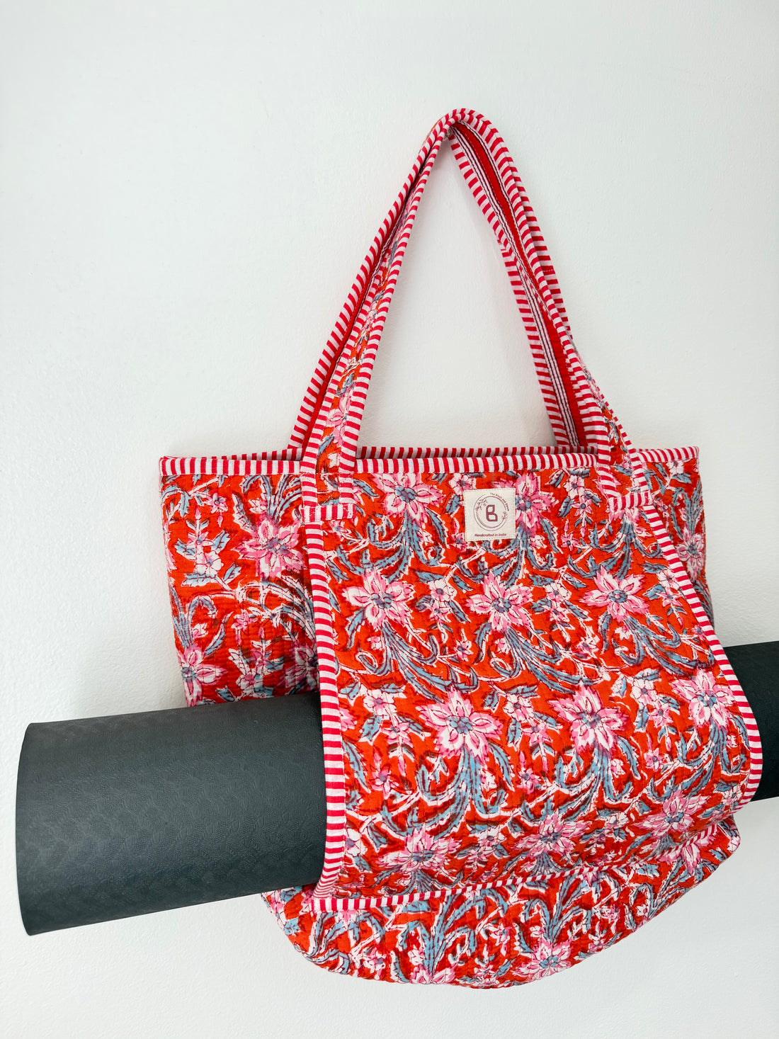 A red fabric tote bag with a pink and blue floral pattern, featuring red and white striped handles and trim, displayed against a white background.