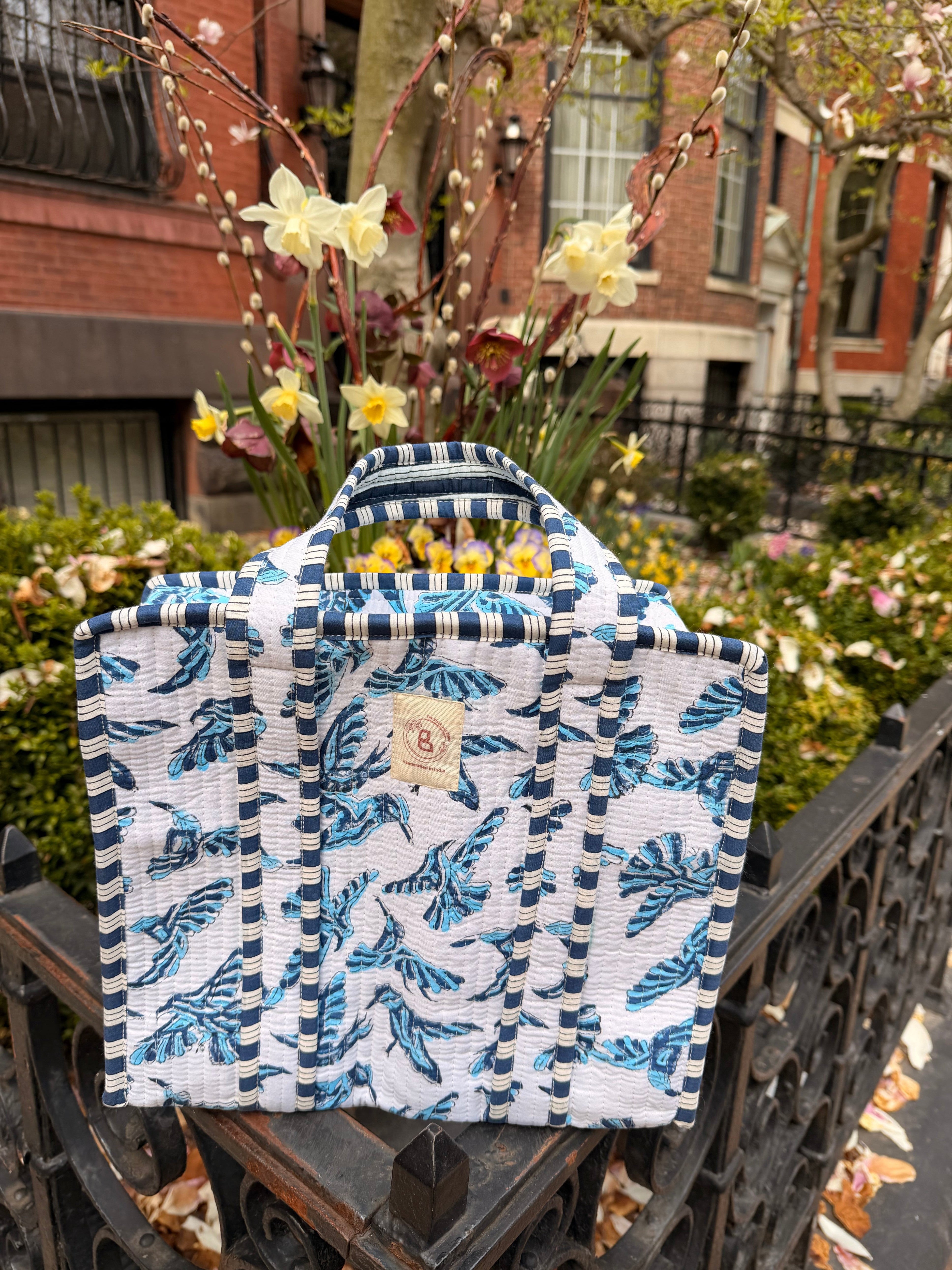 The Block & Bloom blue bird quilted mini tote with hand block-printed pattern and striped handles in a garden setting.