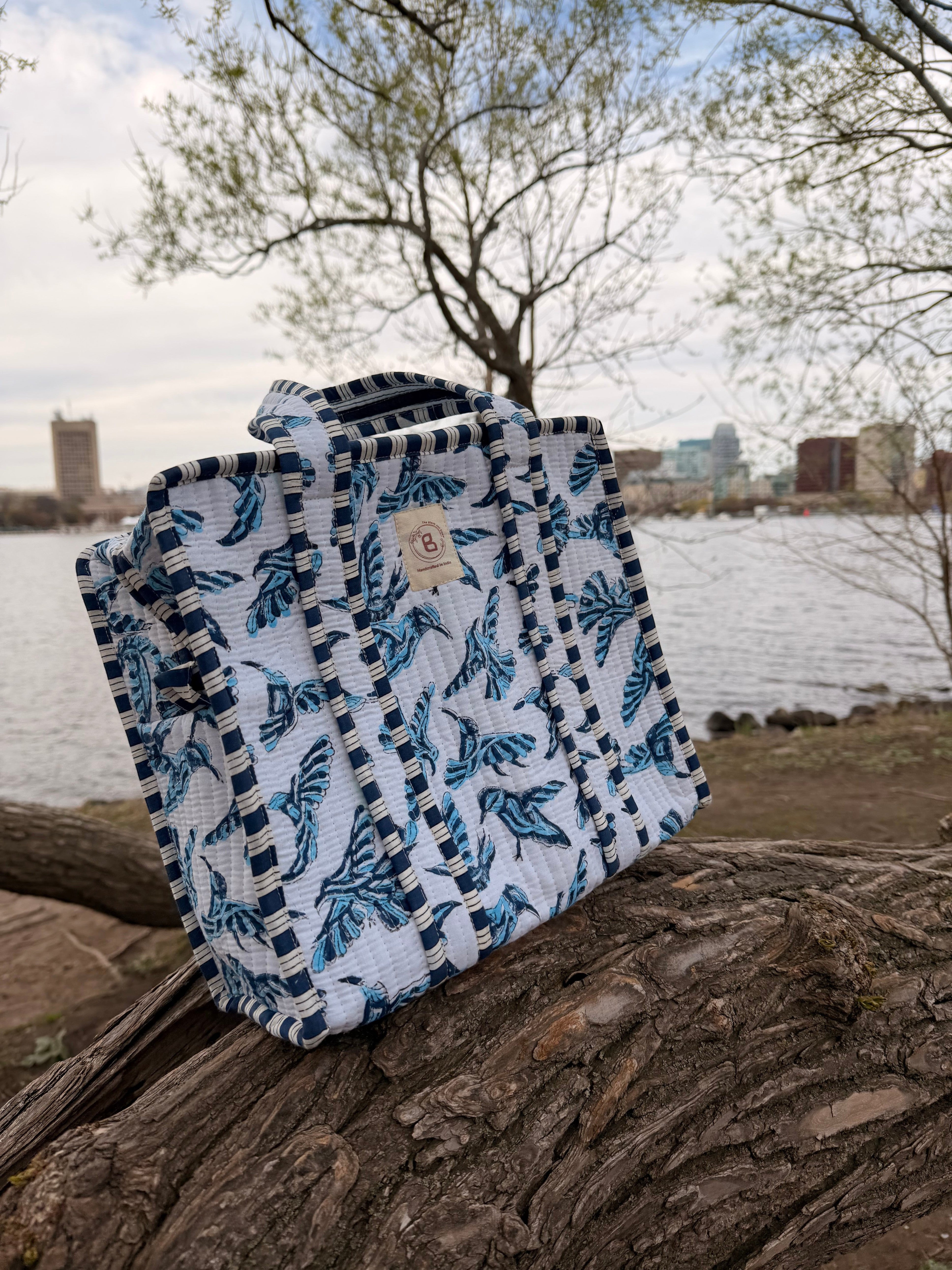 The Block & Bloom quilted mini tote featuring a blue bird block print on white fabric, set on a log by a lake.