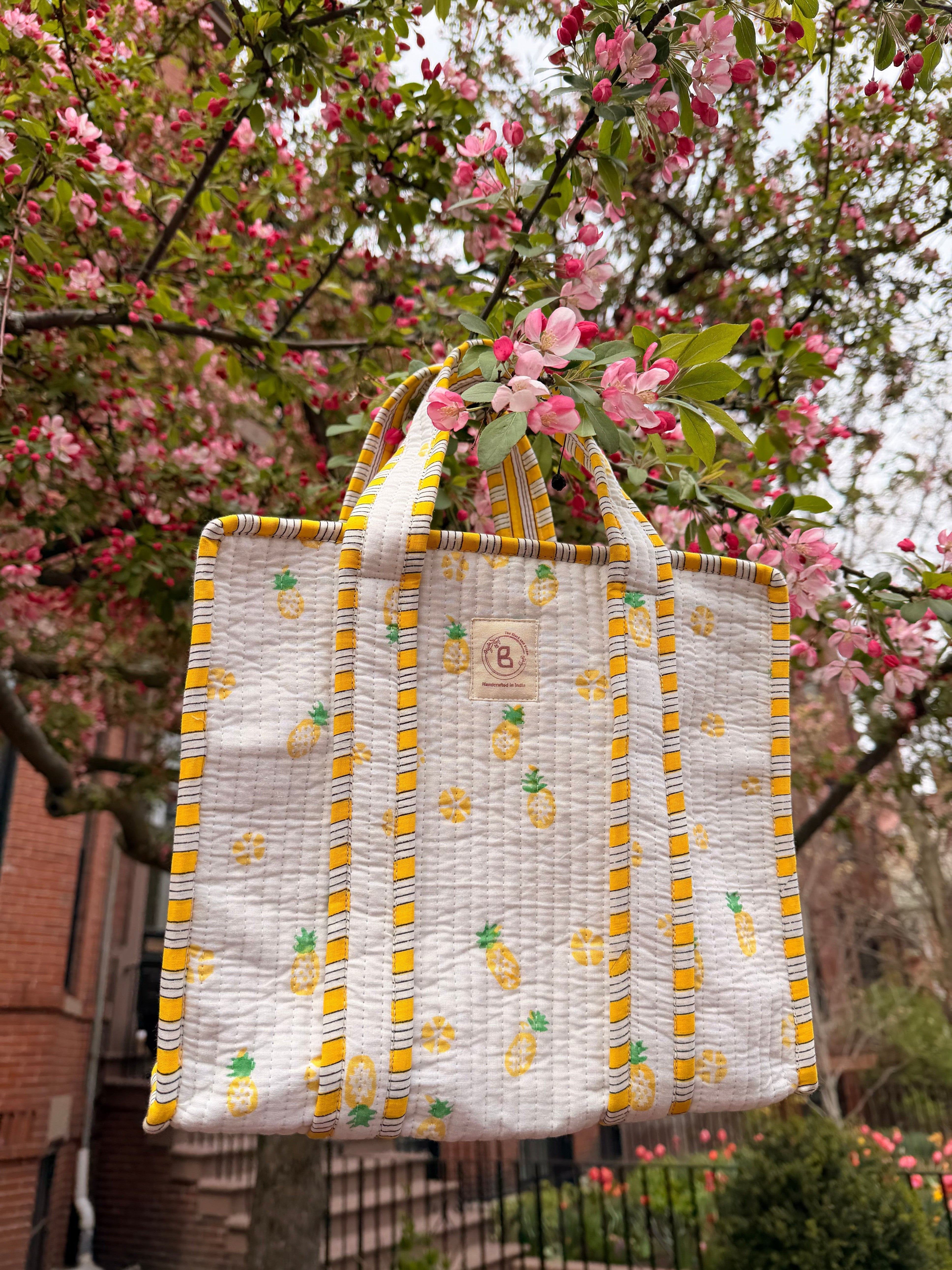 The Block & Bloom quilted mini tote with yellow pineapple print and striped trim hanging in a pink flowering tree.