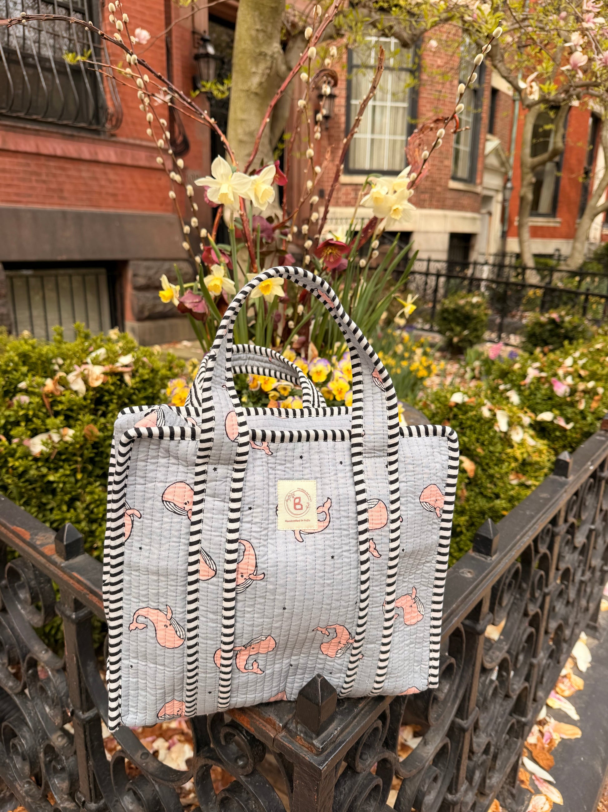 The Block & Bloom quilted mini tote in grey with pink whale print and black-and-white striped trim in a garden setting.