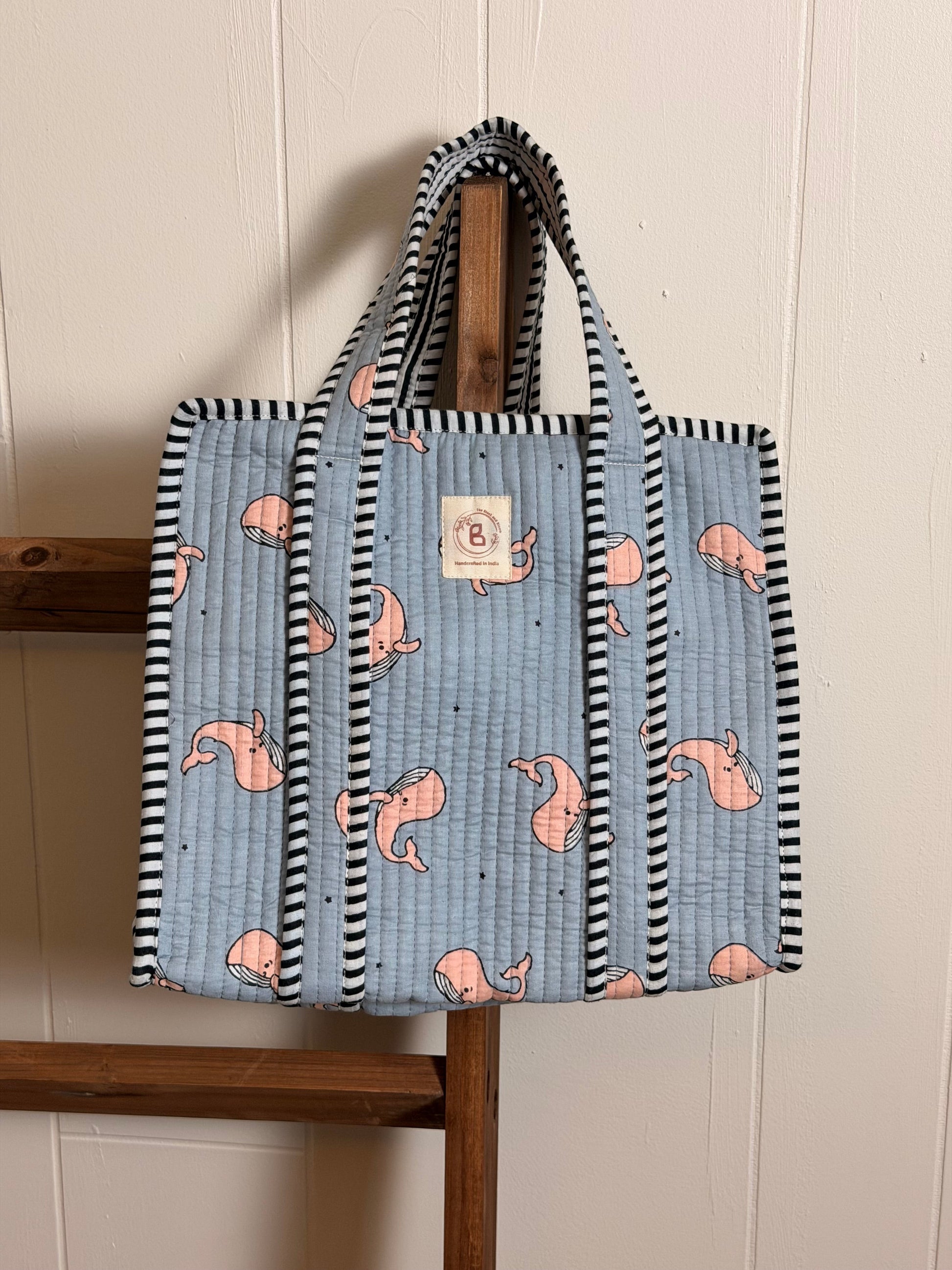 The Block & Bloom quilted mini tote in grey with pink whale print and black-and-white striped trim hanging on a ladder